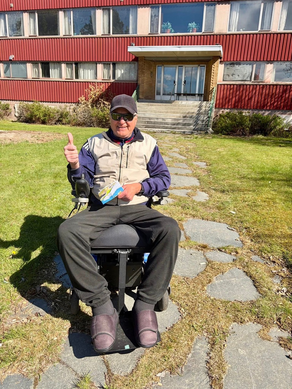 En mann i rullestol sitter utendørs og smiler og gir tommel opp.