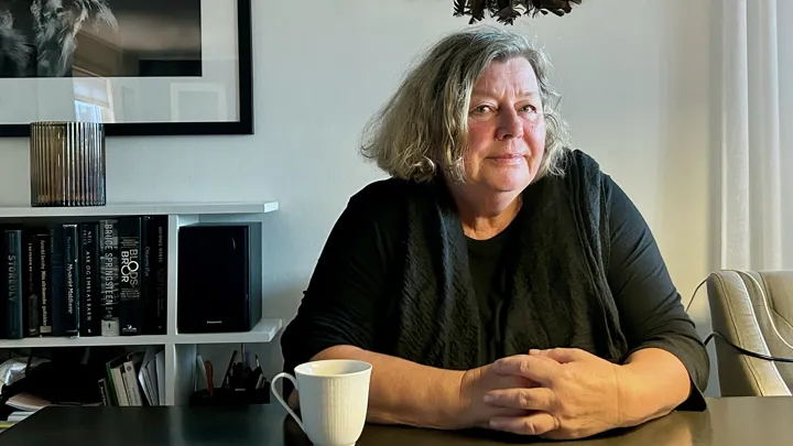 En kvinne sitter ved et bord, og ser skrått inn i kameraet.