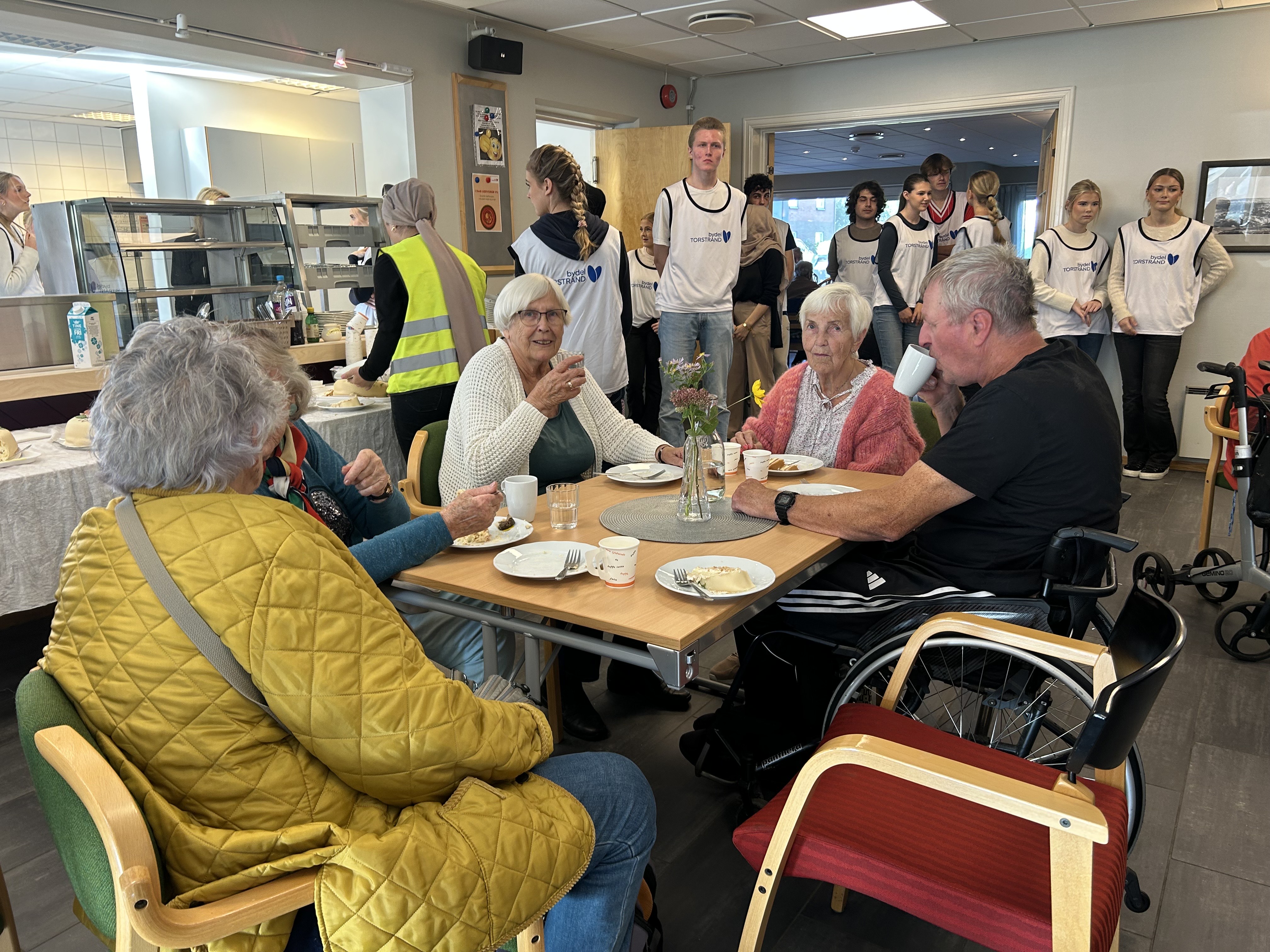 Tre voksne mennesker sitter rundt et bord og drikker kaffe og spiser kake.