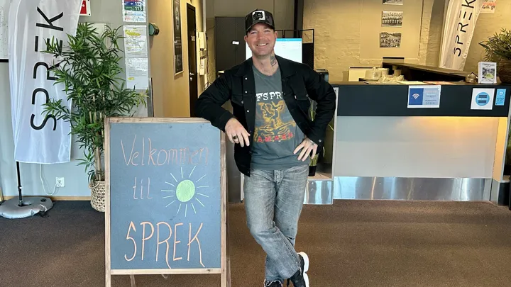 En mann lener seg på et skilt, og smiler mot kameraet.